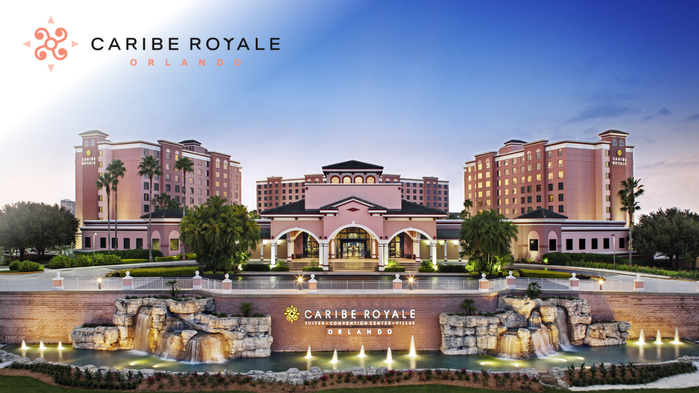 A grand resort complex labeled Caribe Royale Orlando is shown with multiple pink and tan hotel towers surrounding a central entrance. The scene is set at dusk with a dramatic sky, warm architectural lighting, and a large cascading fountain in the foreground. Palm trees and manicured landscaping frame the water features and pathways.